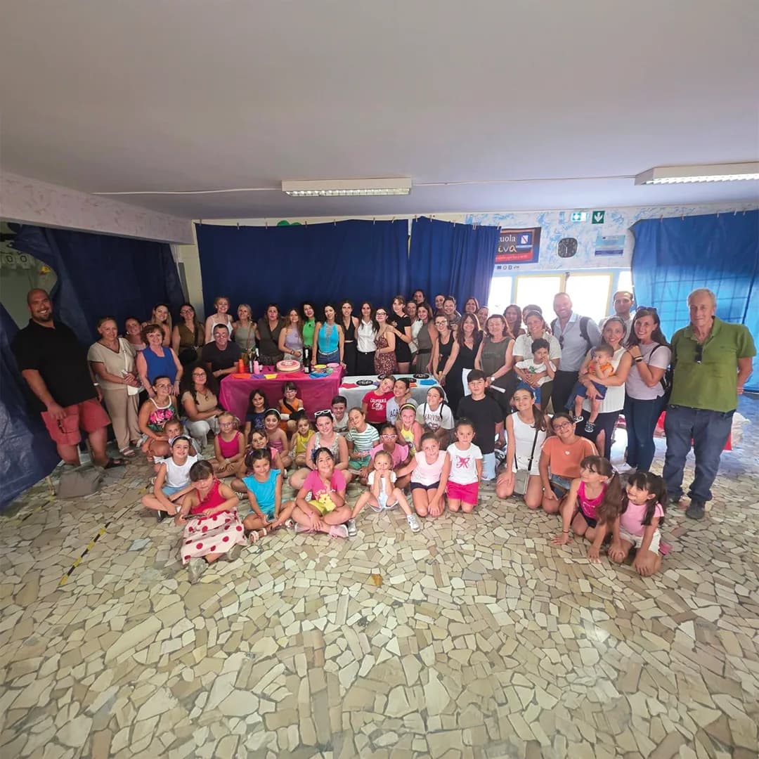 Foto di gruppo al villaggio estivo: bambine, bambini, genitori e volontarie attorno a un tavolo con la torta
